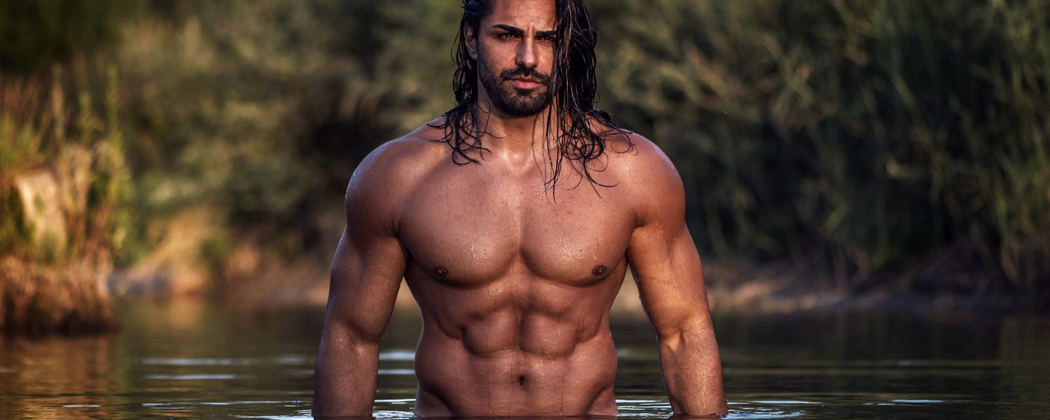 A shirtless, masculine, muscular man standing waist deep in a river, displaying his athletic torso, shoulder length dark hair and chiseled facial feature. 