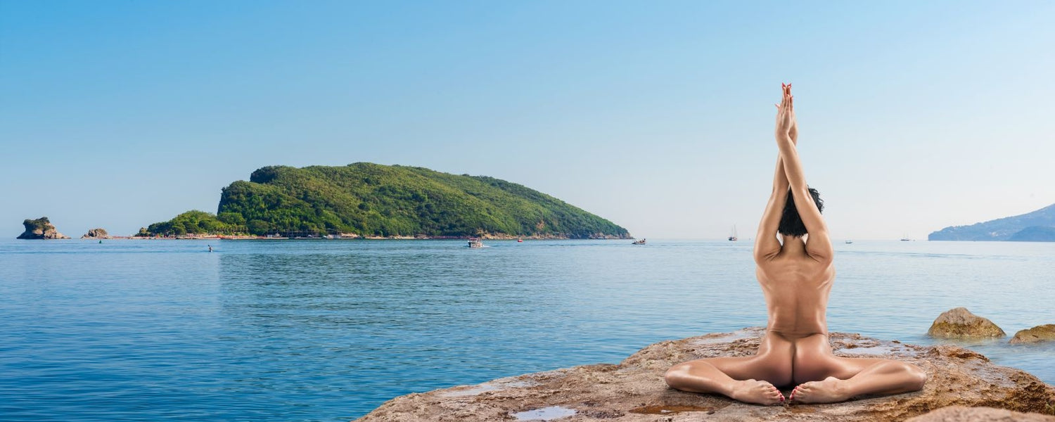 Artistic lifestyle photo of nude yoga model doing extreme butterfly stretch on a rock while overlooking a bay.