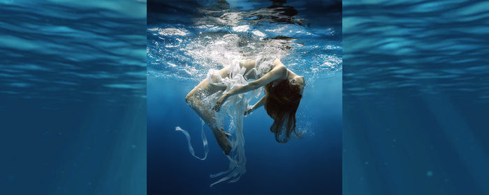 A woman submerged in water, surround by white flowing materials, arch backwards in an elegant and surreal pose.