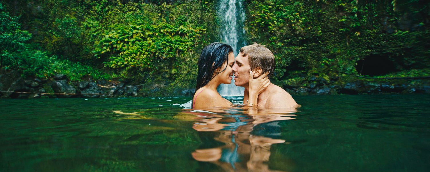 The heads and shoulders of a young couple of a man and a woman are visible as they embrace intimately, their faces touching, bathing in the lush green pool of a waterfall. 